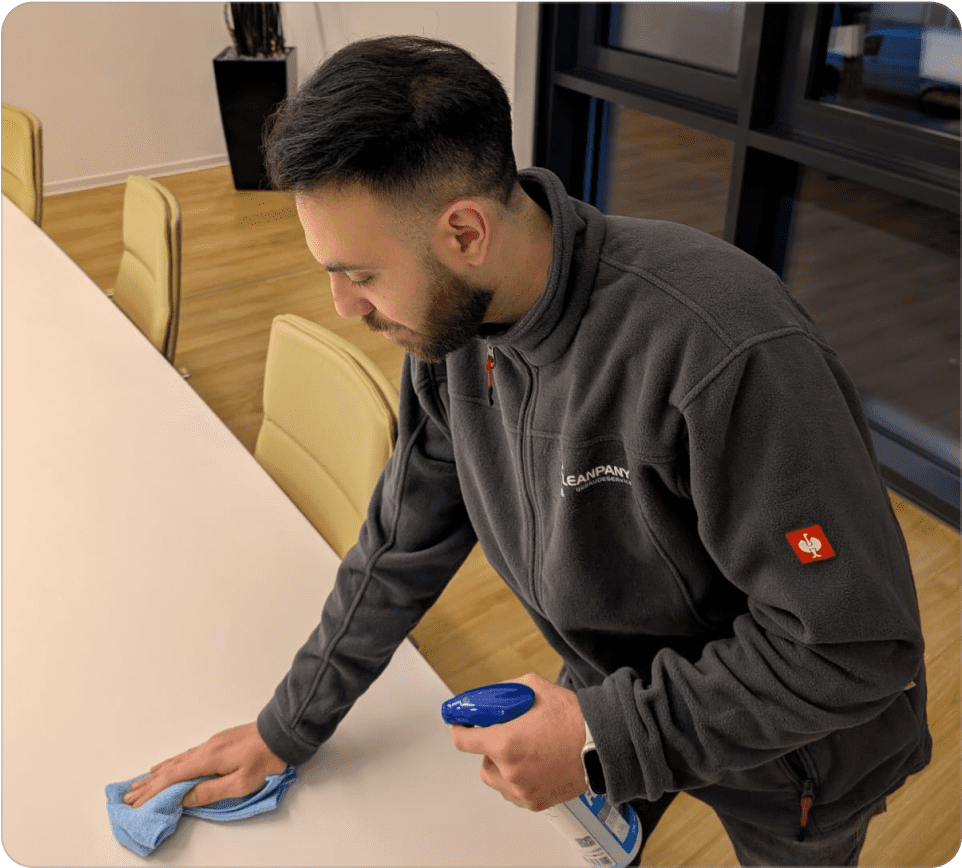 Mitarbeiter reinigt einen Konferenztisch mit Reinigungstuch und Reinigungsmittel.